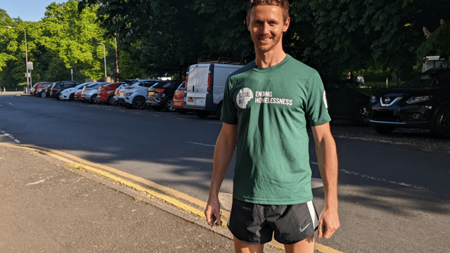 A man in a green tshirt and black running shorts stands on the pavement smiling