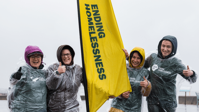 Four women in raincoats and clear ponchos smile and give thumbs up in very wet weather. They have a tall yellow banner between them that reads 'ending homelessness'