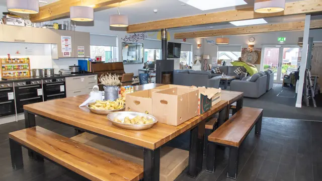 A huge communal kitchen dining table with boxes and snacks on it, looking on to a living room