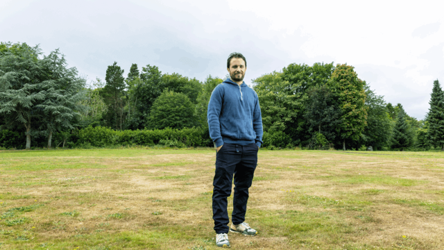 A man in a blue sweatshirt stands in a field with trees
